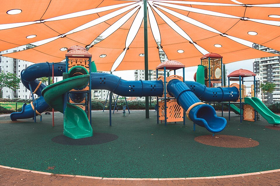 מתקני מגלשה וטיפוס בגן שעשועים ציבורי עם ריצפה רכה וצל
